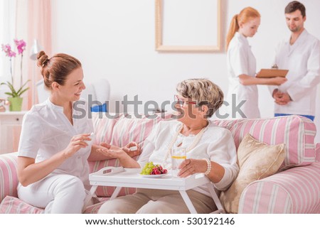 Elderly lady sitting on a couch with a tray and a young nurse giving her medicines in private clinic