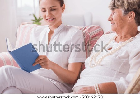 Elegant elderly lady and young attractive nurse, reading book with blue cover