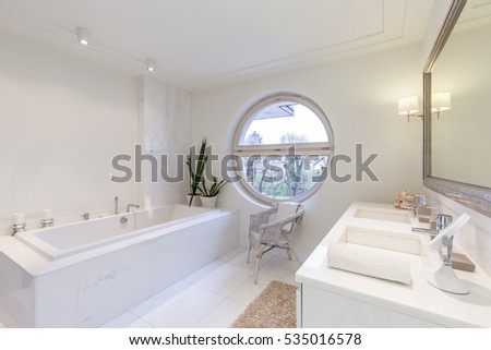 Bright minimalist bathroom with round window, bath, two sinks and large framed mirror
