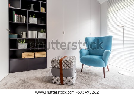 Modern interior with cozy reading corner with black bookcase, blue armchair, pouf and big window with blinds