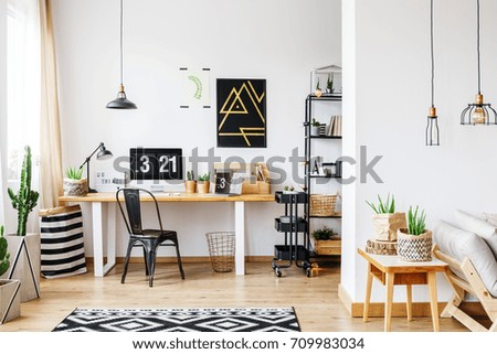 Trendy room with workspace for a freelancer with white wall, desk, black vintage chair, industrial lamps, poster and wooden sofa in bright open living room