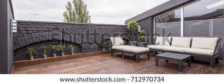 Panorama view of modern rooftop terrace with dark wood deck flooring, plants, brick fence and black garden furniture