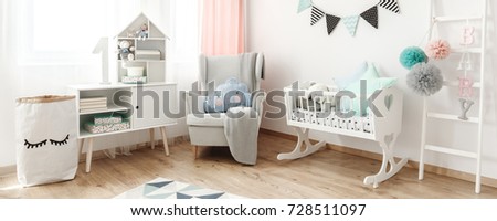 Bright armchair with blanket and cushion standing next to white wooden cradle in pastel baby room
