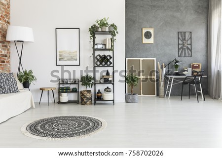 Round patterned carpet in spacious living room with plants, poster and work area against grey wall