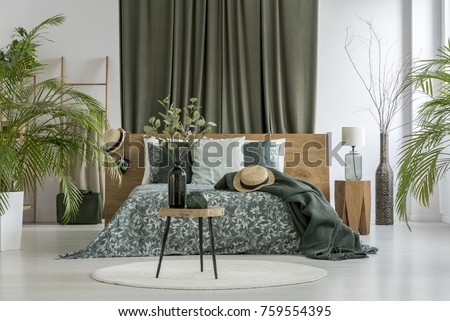 Table with plant on white round carpet and green curtain behind king-size bed with hat in bedroom with plants