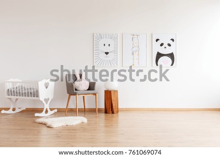 Drawings on white wall above grey chair with pillow and wooden stool in baby\'s room with white crib and rug