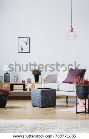 Vase and candle on grey pouf next to wooden cupboard and couch with purple pillow in living room interior with heathers
