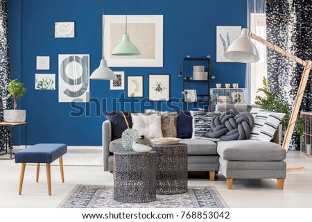 Designer metal tables in living room with navy blue stool near grey corner sofa and lamp