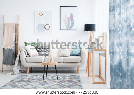 Blue curtain, posters and carpet in living room with blankets on ladder next to sofa with pillows