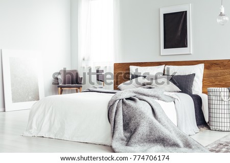 Grey blanket on bed with wooden bedhead in simple bedroom interior with dark poster and chair under window