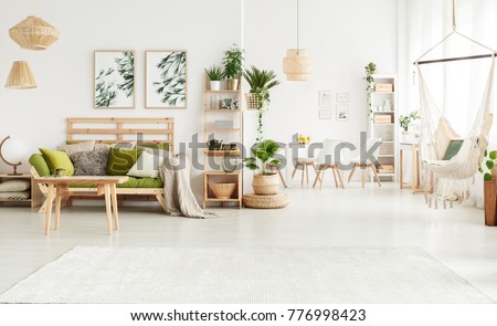 Plant on pouf and leaves posters in bright open space interior with green couch, hammock and white chairs at a table