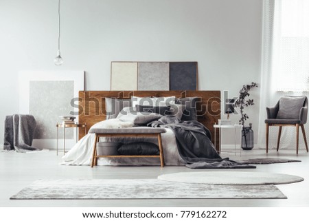 Simple painting on wooden bedhead of bed with pillows in grey bedroom interior with rugs, bench and chair