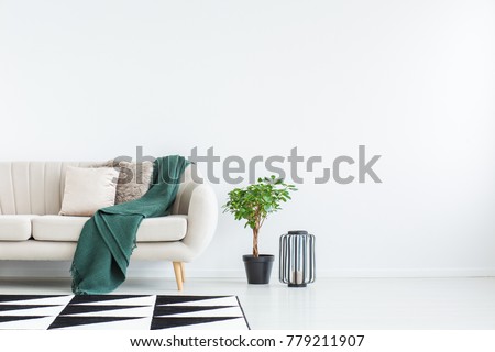 Beige sofa with green blanket and cushions standing against white, empty wall in minimalistic living room
