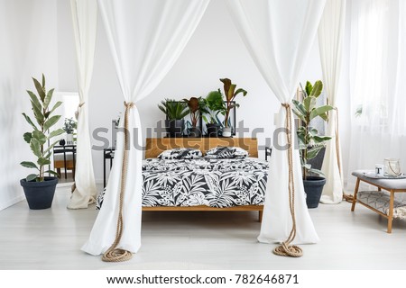 Plants on wooden bedhead of bed with patterned bedding and white drapes in bright bedroom interior with grey bench