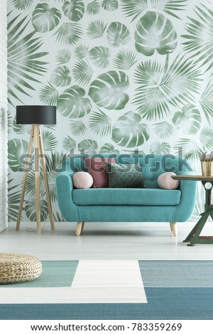 Blue sofa and wooden lamp against white wallpaper with green monstera leaves in natural living room interior