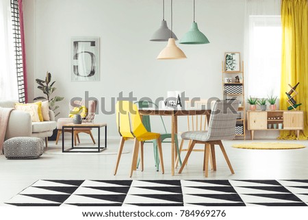 Geometric carpet in cozy living room interior with pastel lamps above wooden table and yellow, mint and grey chair against a wall with poster