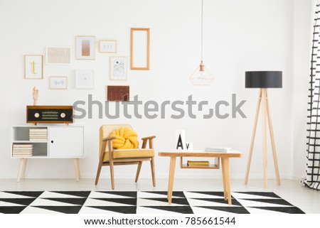 Wooden table on black and white carpet near lamp and vintage armchair with yellow pillow in bright apartment interior with radio on cupboard against wall with posters