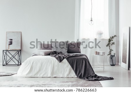 Simple, gray and white bedroom interior with blanket and pillows on king size bed, bright window and posters