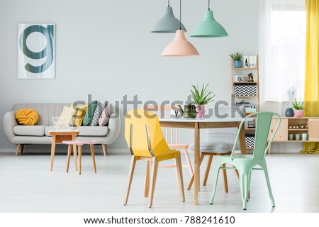 Modern colorful chairs at dining table under pastel lamps in living room interior with pillows on settee against wall with poster