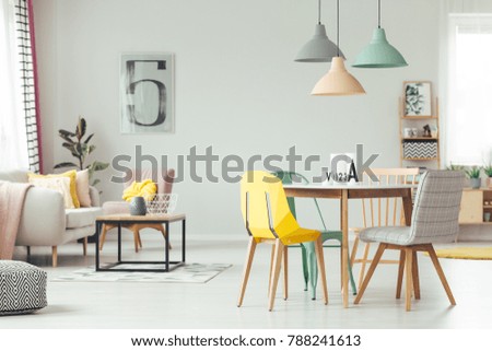 Colorful lamps above wooden table and chairs in yellow living room interior with poster on the wall and pink armchair next to settee