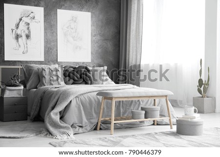 Wooden bench and boxes in monochromatic bedroom interior with drawings on concrete wall above bed