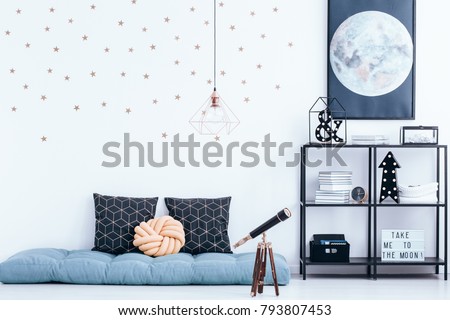 Telescope next to blue mattress with cushions in kid's bedroom interior with stars stickers and moon poster on white wall above shelves