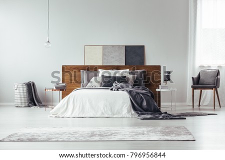 Dark bedsheets on bed with wooden bedhead and beige rug in bright bedroom interior with lamp on table next to armchair