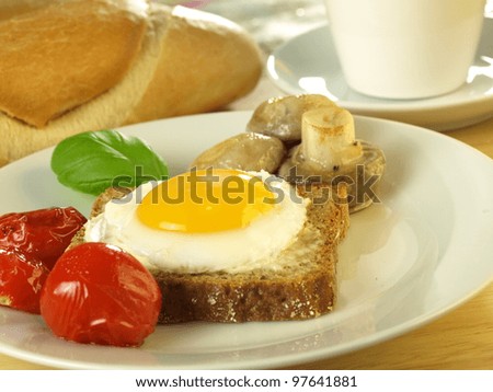 Sunny side up egg on a slice of bread with tomatoes and mushrooms on a plate with a French loaf and tea cup in the background