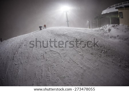 Two Snowboarders Strap in to their boards at the top of a chilly mountain at night. It looks like a frigid frosty scene at this ski area