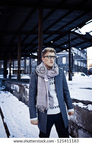 Stylish man in a jacket, sweater, scarf and glasses on the background of the industrial