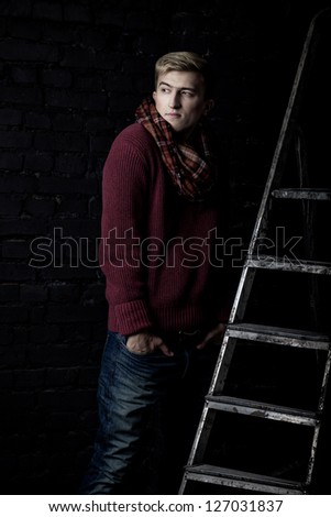 Elegant man in a sweater, jeans and a scarf standing near a ladder against a brick wall
