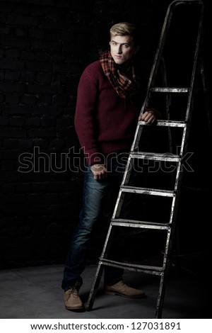 Elegant man in a sweater, jeans and a scarf standing near a ladder against a brick wall