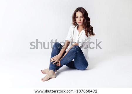 Fashion model sitting on a floor in a blouse and jeans barefoot on a white background
