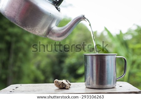 Pour water from aluminium pot  into a stainless steel cup on old wood table With blur green background from a tree soft.
