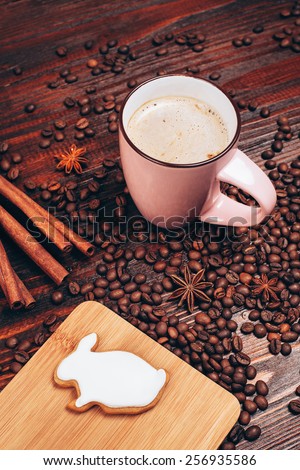Cup of coffee with foam, cinnamon sticks, star anice, coffee beans and wooden board with bunny-shaped easter cookie, standing on the wooden table