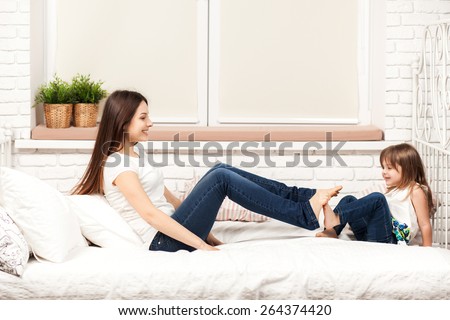young sexy mother spending daytime, having fun with her daughter, lying on the bad with pillows in light room