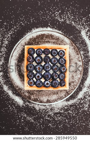 Blueberry tart on a glass saucer on black background and spilling sugar