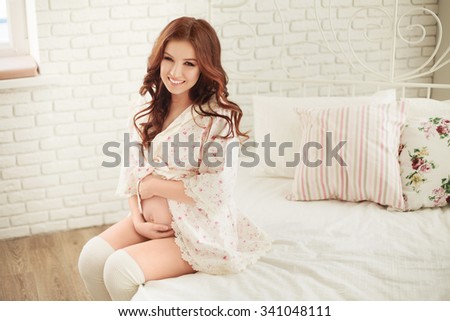 Pregnant Happy smiling Woman sitting on the bed in lace robe and caressing her belly. Mom Expecting Baby. Pregnant Woman Belly. Pregnancy. Beautiful Pregnant Woman. Maternity concept. Baby Shower