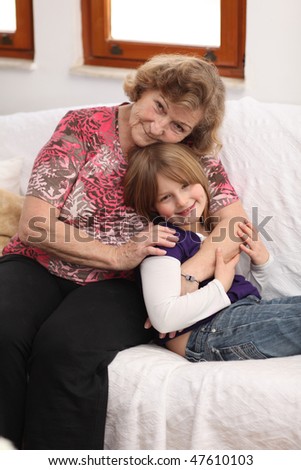 Young girl with blonde hair, purple dress and blue jeans lying on sofa embraced by her grandmother, smiling to the camera.