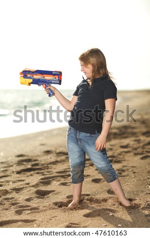 Happy blonde young girl with a colorful toy gun on the beach looking to the sea, back light, selective focus on gun