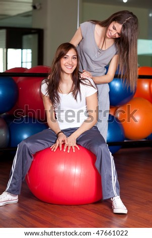 Two happy young women sitting on a red ball in the gym smiling to the camera