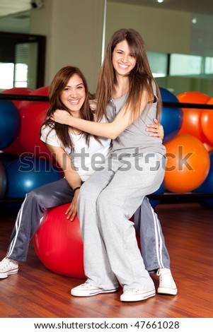 Two happy young women in the gym sitting on a red ball smiling to the camera