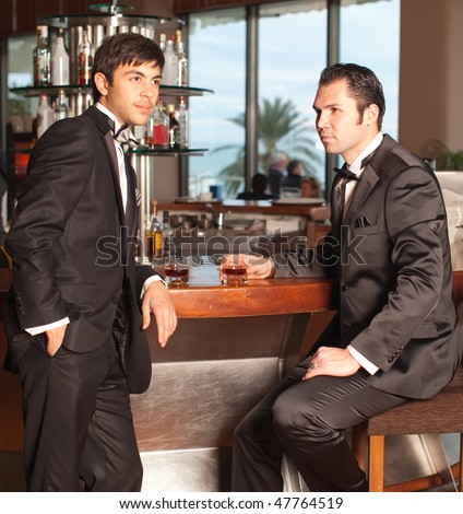 Two handsome young men in a black tuxedo at a round bar drinking whiskey, palm tree in the background