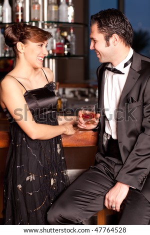 Young couple at a bar, men in black tuxedo holding whiskey in his hand, woman with hairstyle in black dress smiling