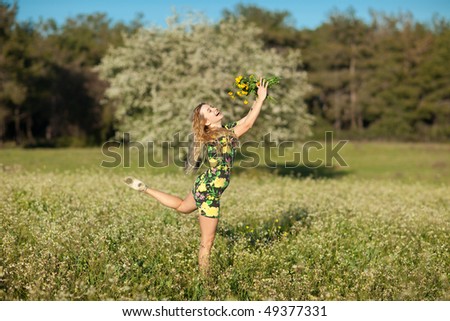 Beautiful young blonde woman jumping in blooming meadow in spring, bunch of yellow flowers in hand; shallow depth of field, trees in background