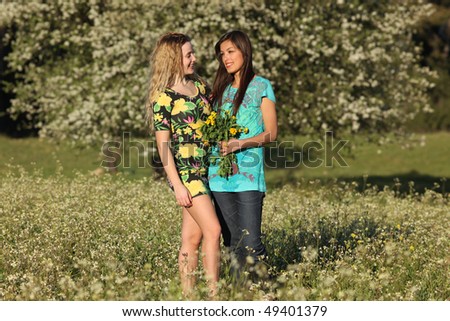Two beautiful young women standing in blooming meadow in spring, smiling, trees in background; shallow depth of field