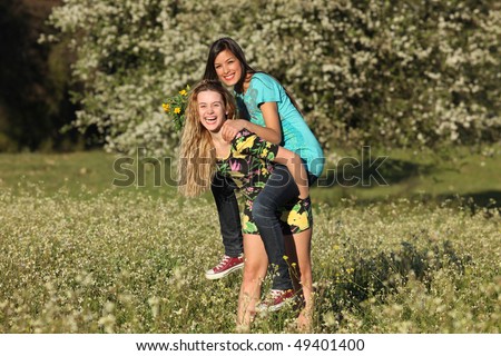 Two beautiful young women standing in blooming meadow in spring, giving piggyback, bunch of yellow flowers in hand, smiling, tree in background; shallow depth of field
