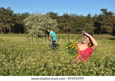 Two beautiful young women standing in blooming meadow in spring, smiling, blue sky and trees in background; shallow depth of field