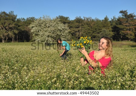 Two beautiful young women standing in blooming meadow in spring, smiling, blue sky and trees in background; shallow depth of field