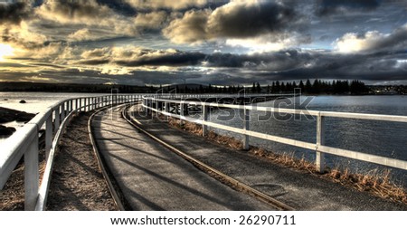 A train track leading the way across a jetty to the mainland. Taken at sunset with a brooding sky.